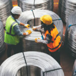 Aerial view of young engineer man and industrial worker woman checking production line and testing quality of steel pipe coils at warehouse section in cable factory. XXXL